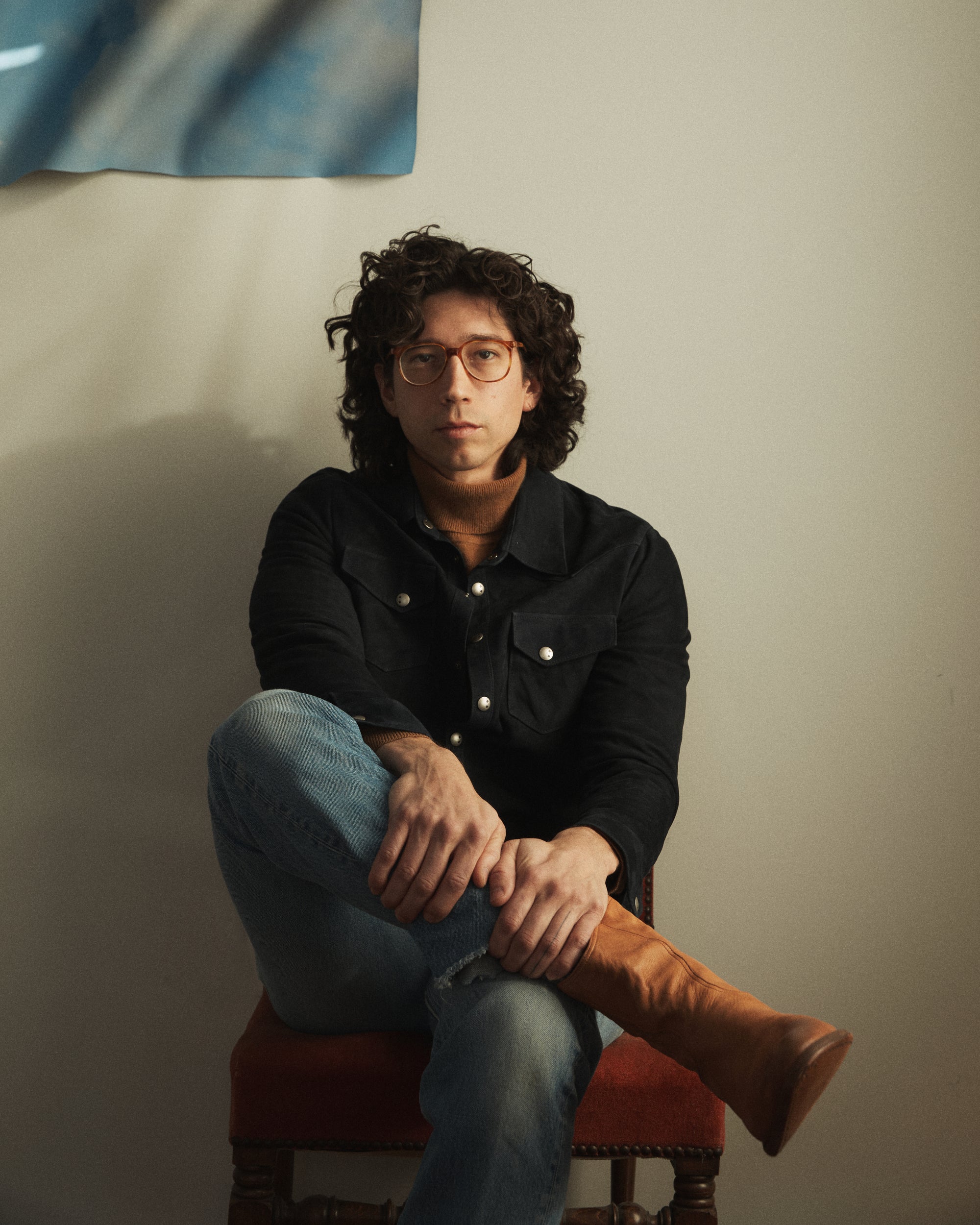 Man in glasses sitting with leg crossed over knee, tan boot wearing navy suede shirt over camel color turtleneck