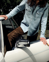 Model wearing The Allen light blue denim shirt in Japanese selvedge cotton, styled with sleeves down and pearl snap front. Posed in front of vintage car.