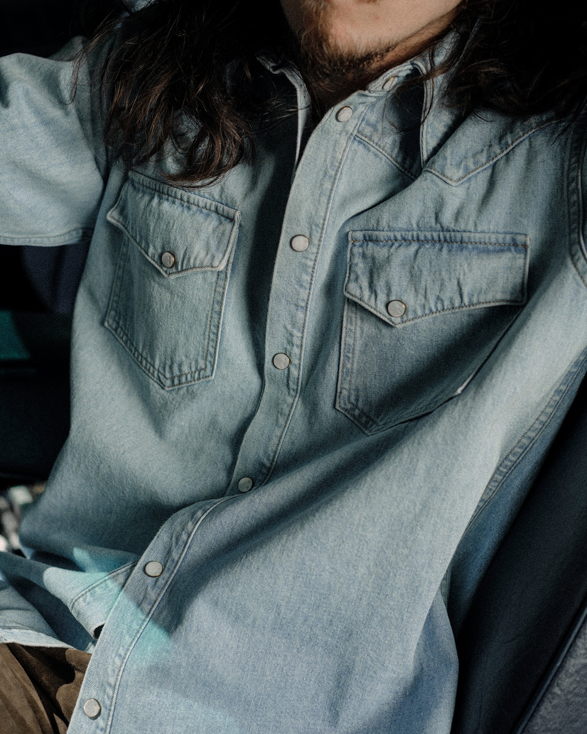 Model wearing The Allen light blue denim shirt in Japanese selvedge cotton, styled with sleeves down and pearl snap front. Posed in front of vintage car.