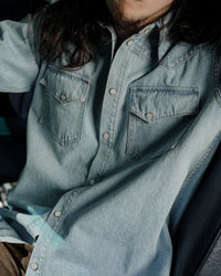 Model wearing The Allen light blue denim shirt in Japanese selvedge cotton, styled with sleeves down and pearl snap front. Posed in front of vintage car.