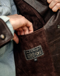 Close-up of a brown leather jacket with a brand label, showing a hand reaching into the pocket.