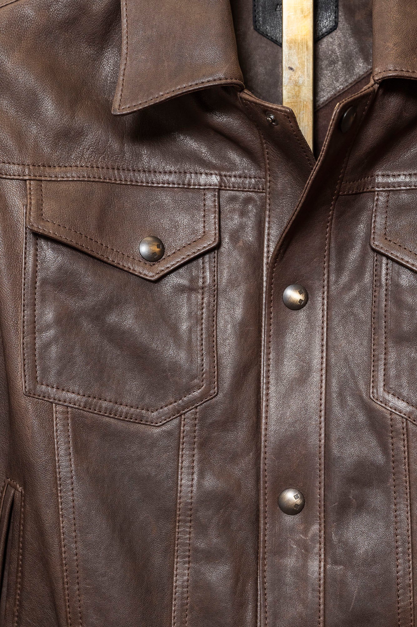 Close-up of a brown leather jacket with buttons and a collar. Softly tailored leather jacket in black lambskin, Savas Denham with structured yet comfortable fit