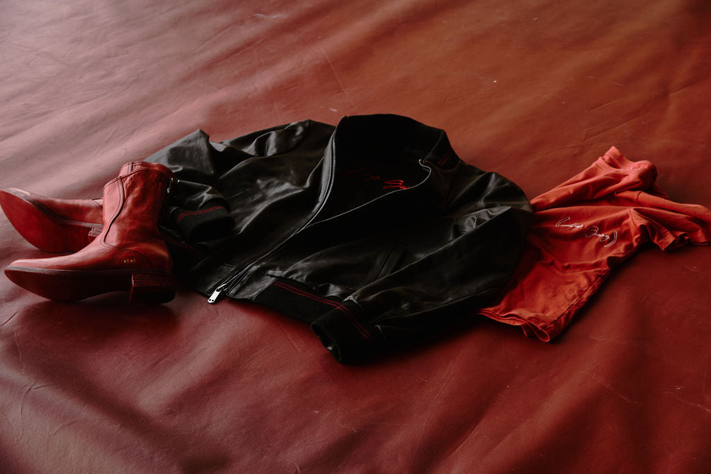 Three Savas (RED) collaborative pieces, red boots, bomber jacket, and red t-shirt on a brown leather background.
