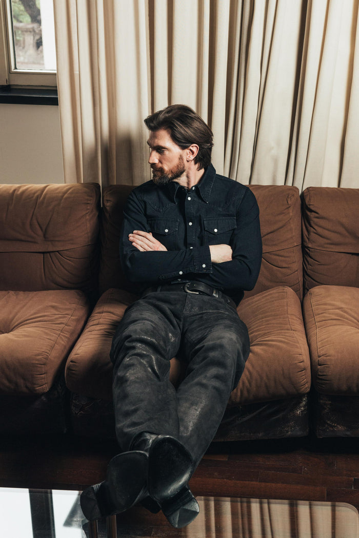 Model on couch wears The Allen selvedge denim shirt in black. Designed by Atelier Savas.  Handmade in LA.