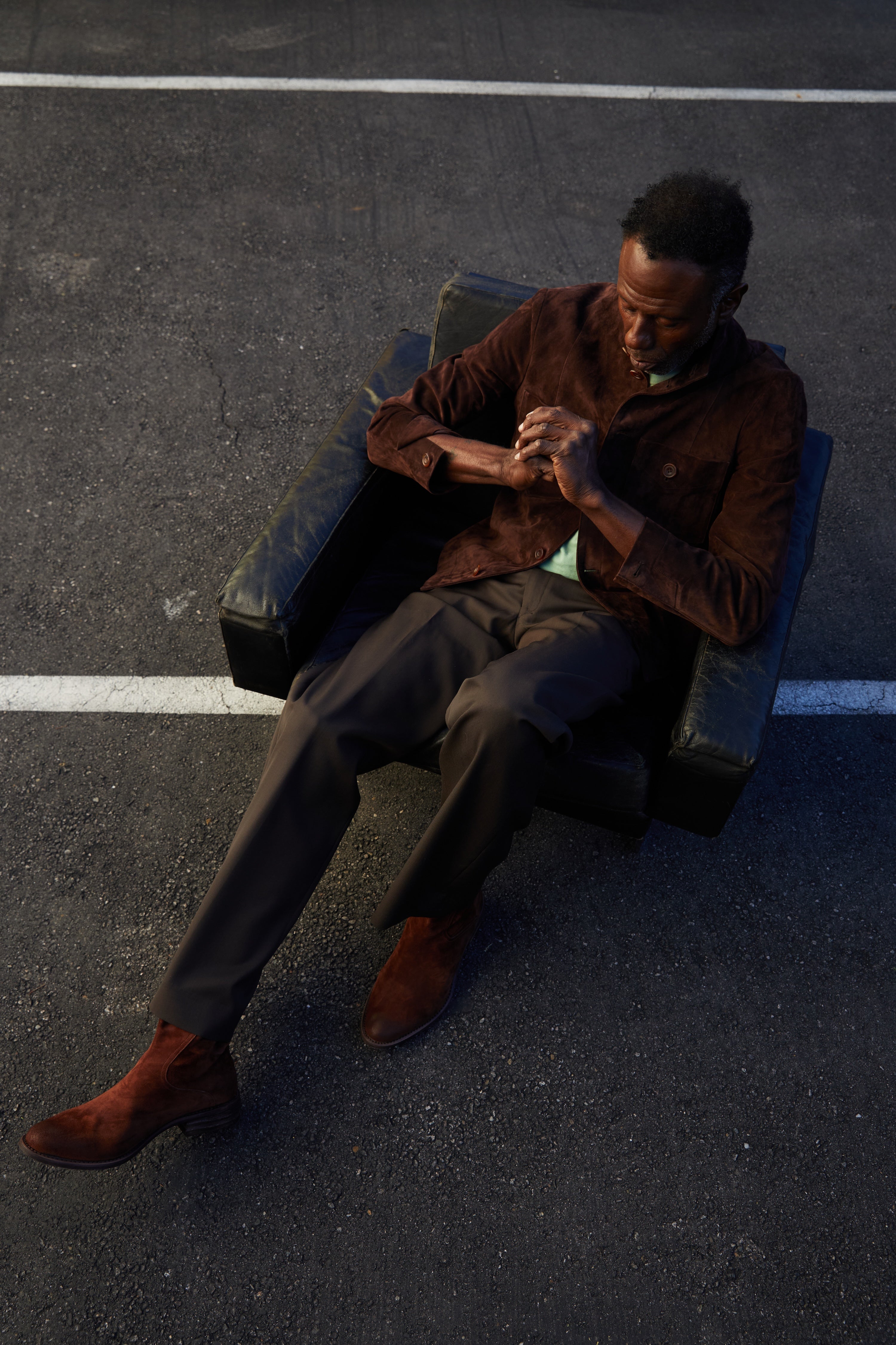 Man sitting on a bench in an urban setting, wearing a brown coat and shoes.