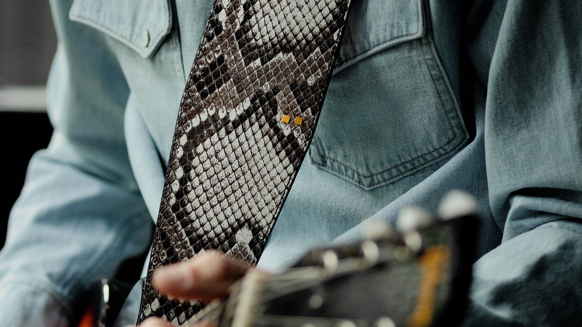 Person playing guitar with a snake skin strap, wearing a denim jacket.