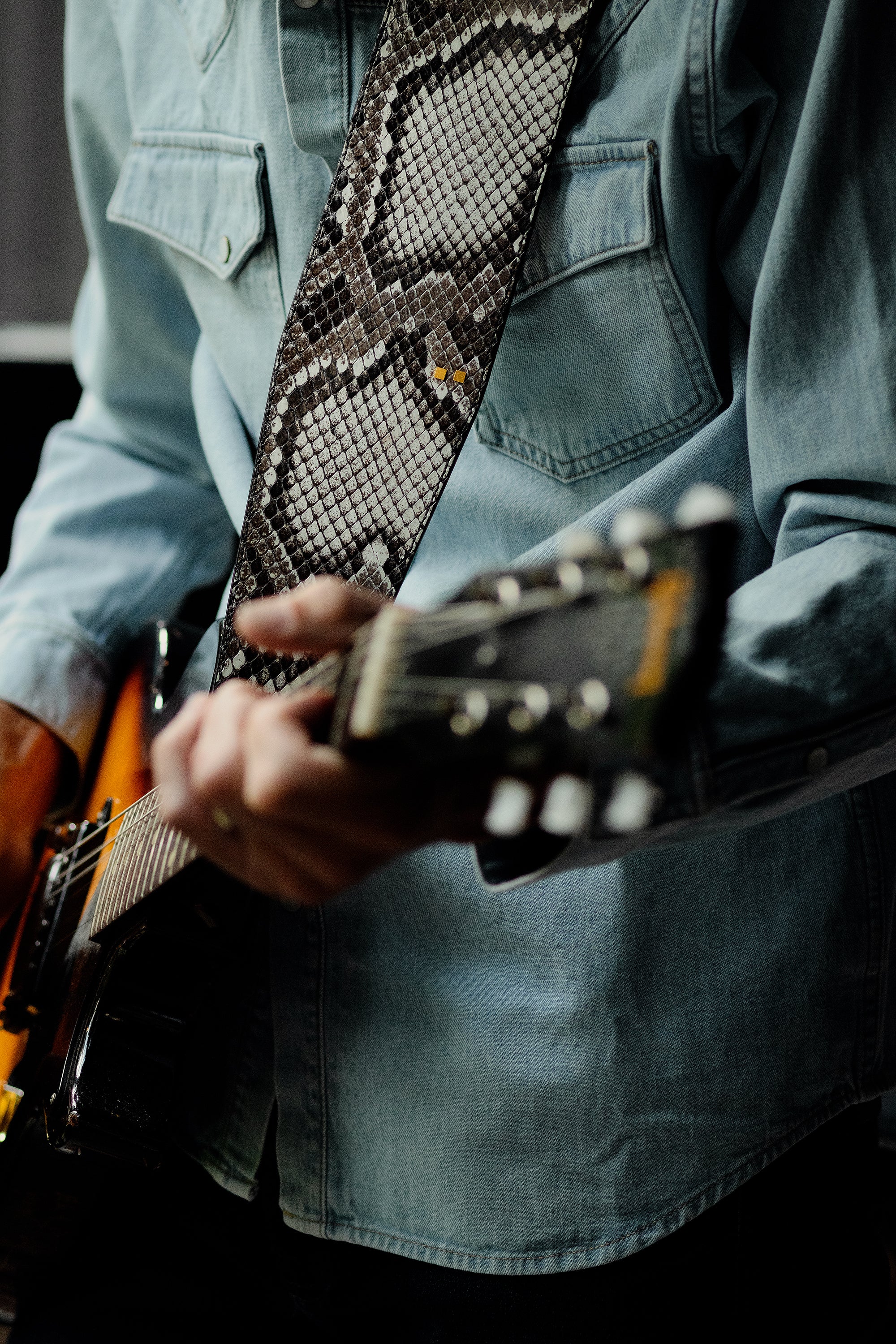 Person playing guitar with a snake skin strap, wearing a denim jacket.