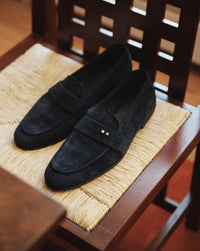 The Coyle sapphire suede loafer resting on a rush-seat chair, showing deep navy tone of Italian calf suede and Two Dots detailing
