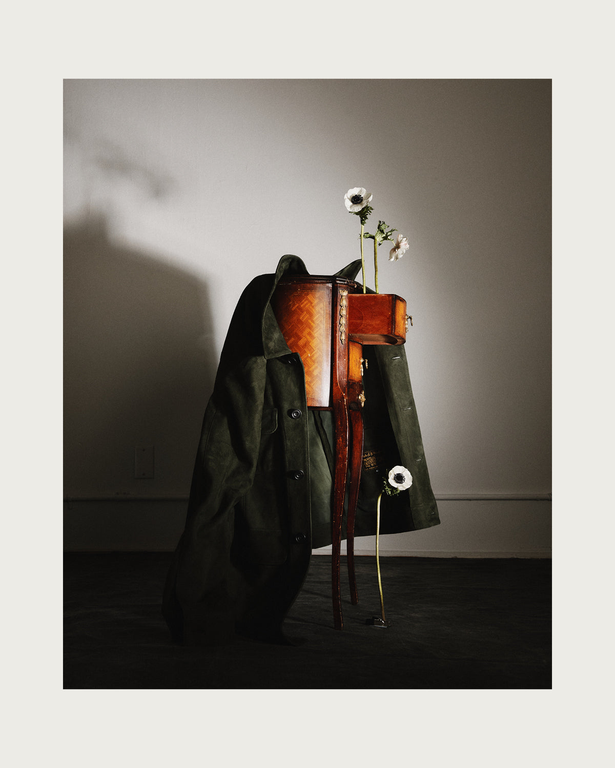 Dark green coat draped over a wooden stand with a vase of flowers on a gray background