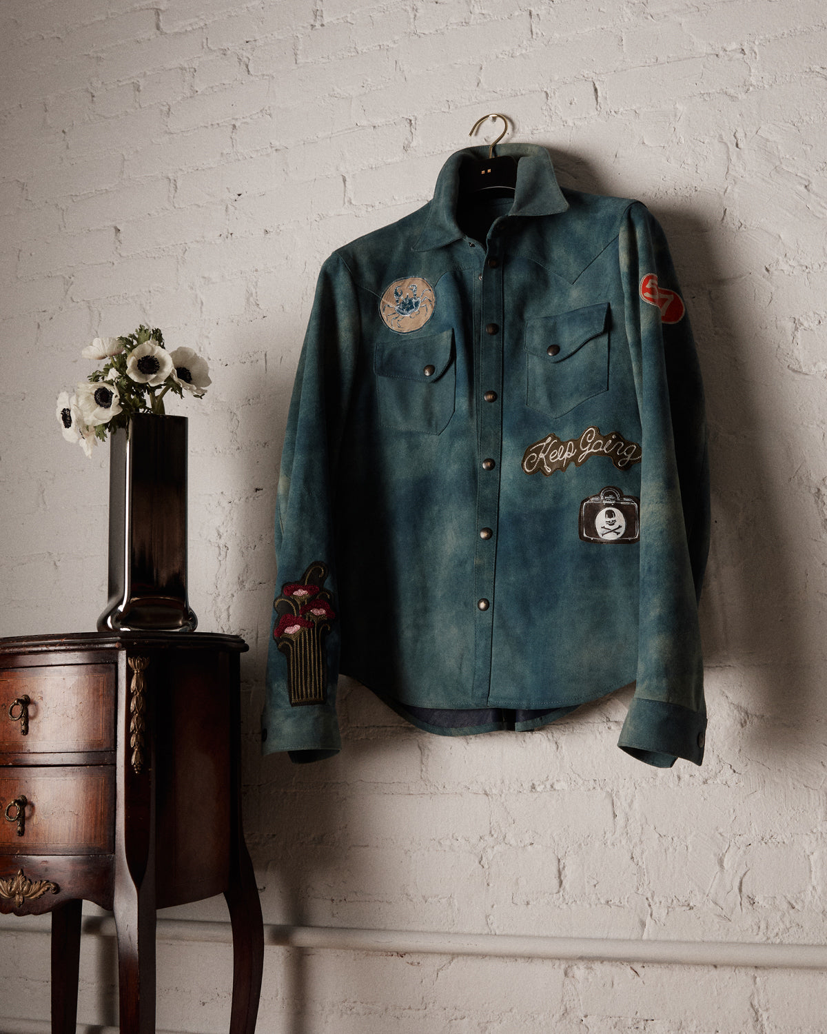 Blue suede shirt with patches hanging on a white brick wall next to a wooden side table with flowers.
