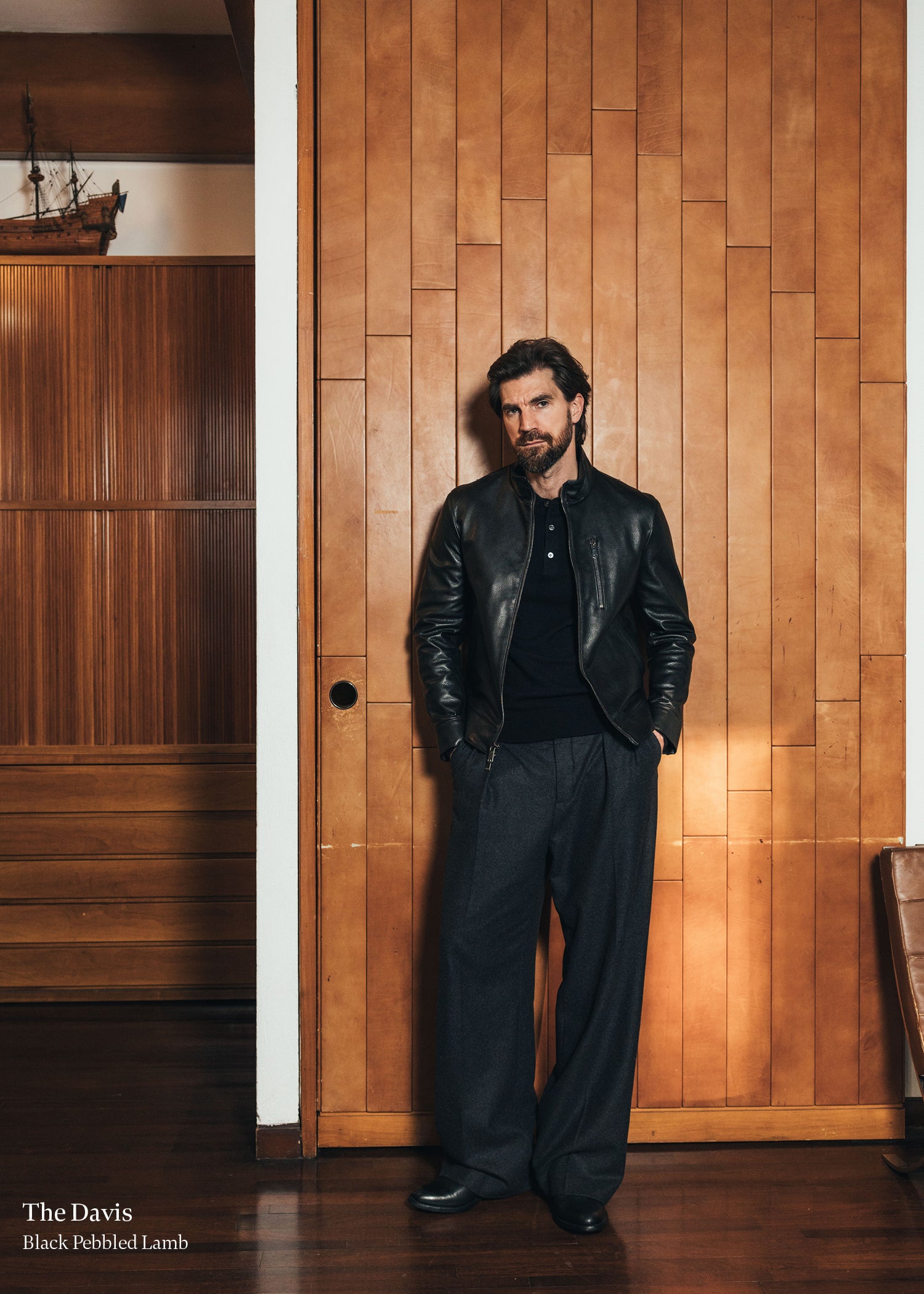 Man in a leather jacket and dark pants standing against a wooden wall.