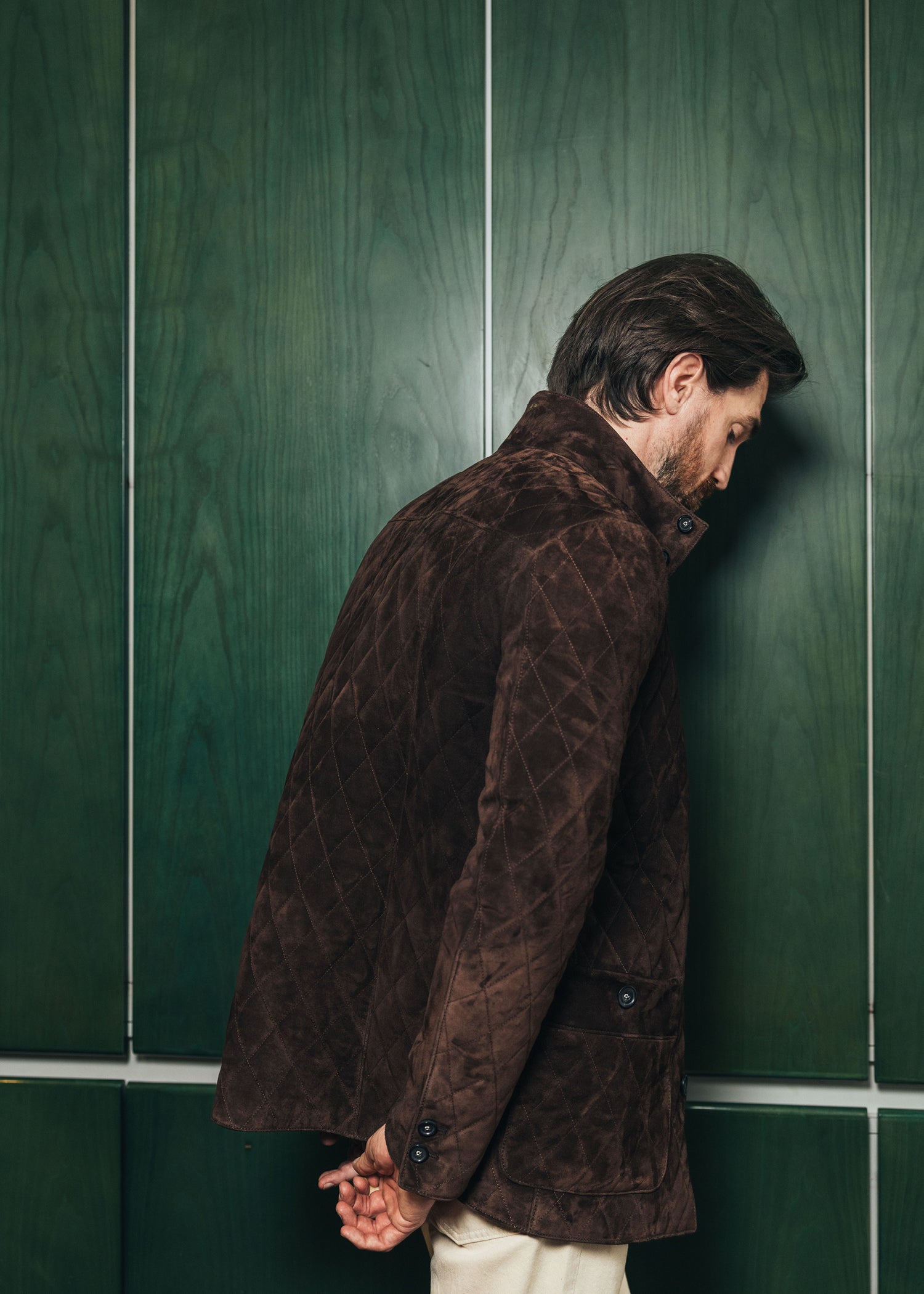 Man in a brown quilted jacket leaning against a green tiled wall.