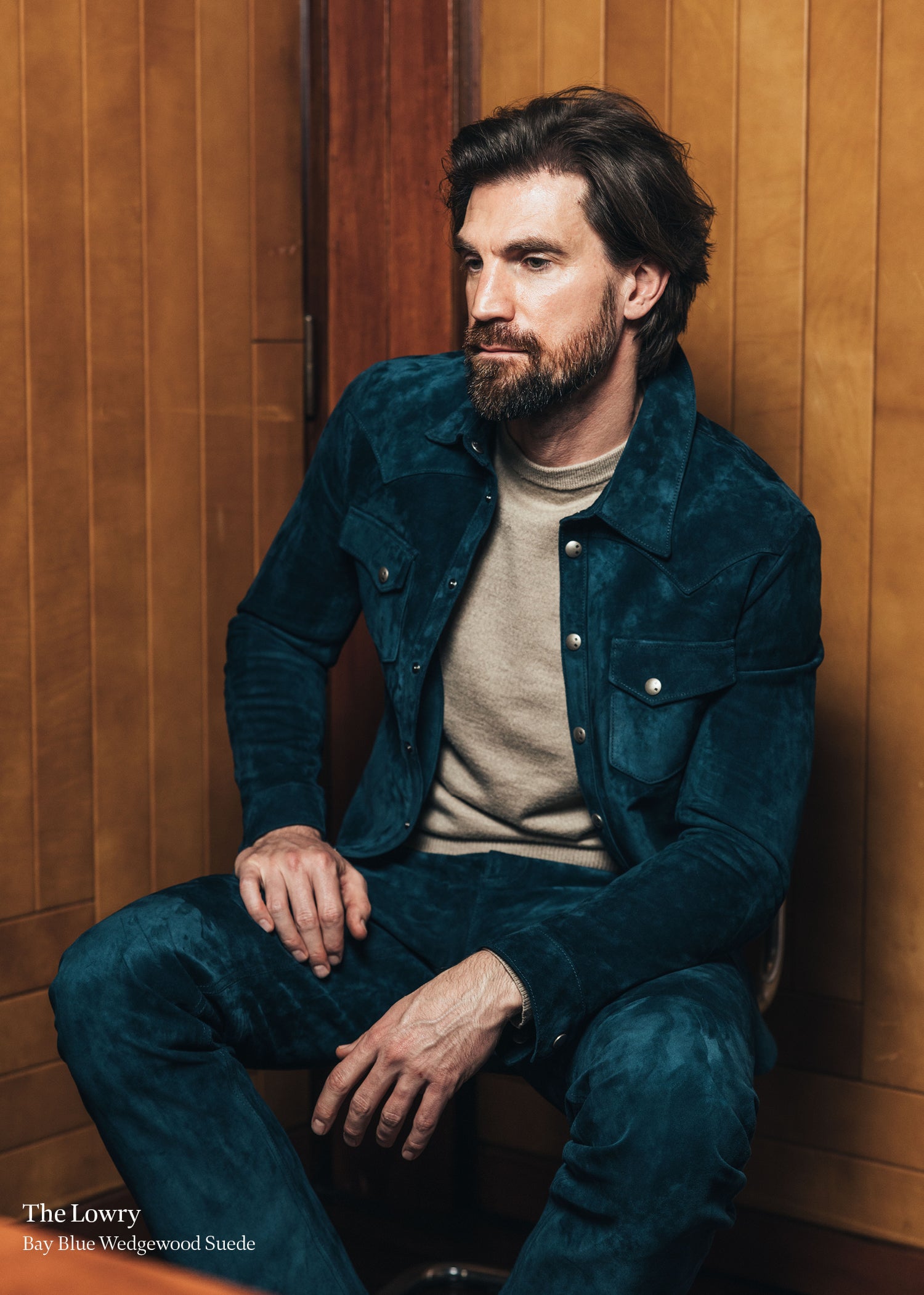Man wearing a blue jacket and beige sweater against a wooden paneled wall.