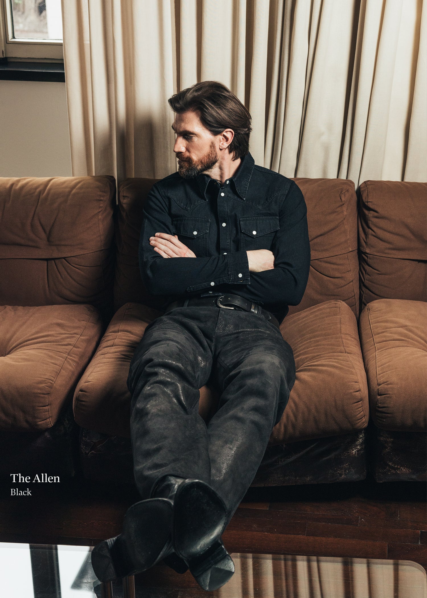 Man sitting on a brown couch wearing a black shirt and jeans, with curtains in the background.