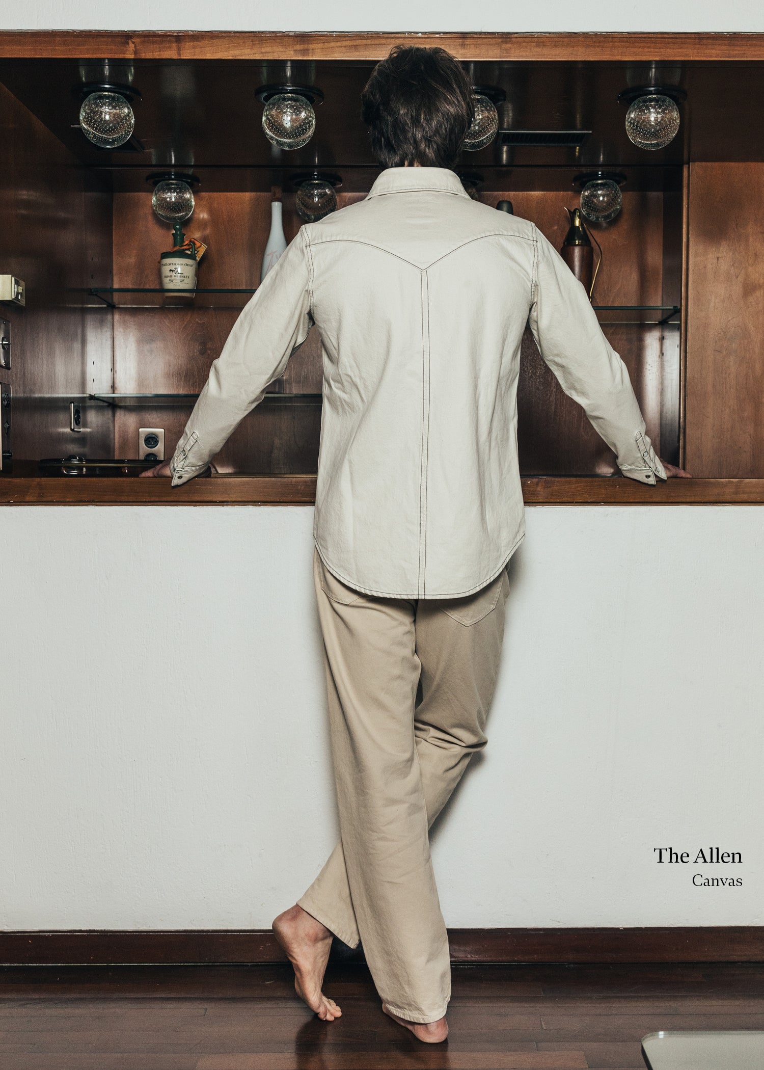 Person wearing a beige jacket and pants standing in front of a wooden shelf.
