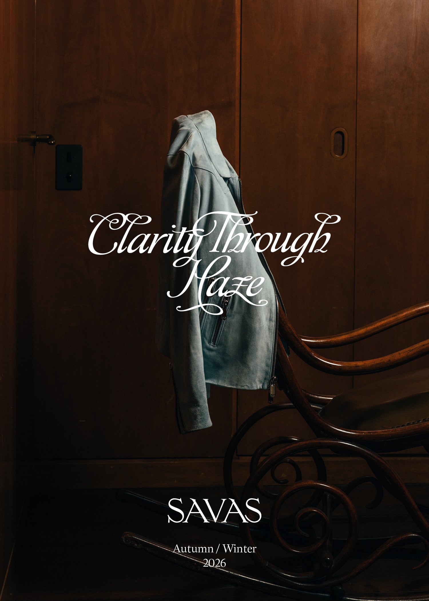 Gray coat hanging on a wooden door with text 'Clarity Through Haze' and 'SAVAS Autumn/Winter 2026'.