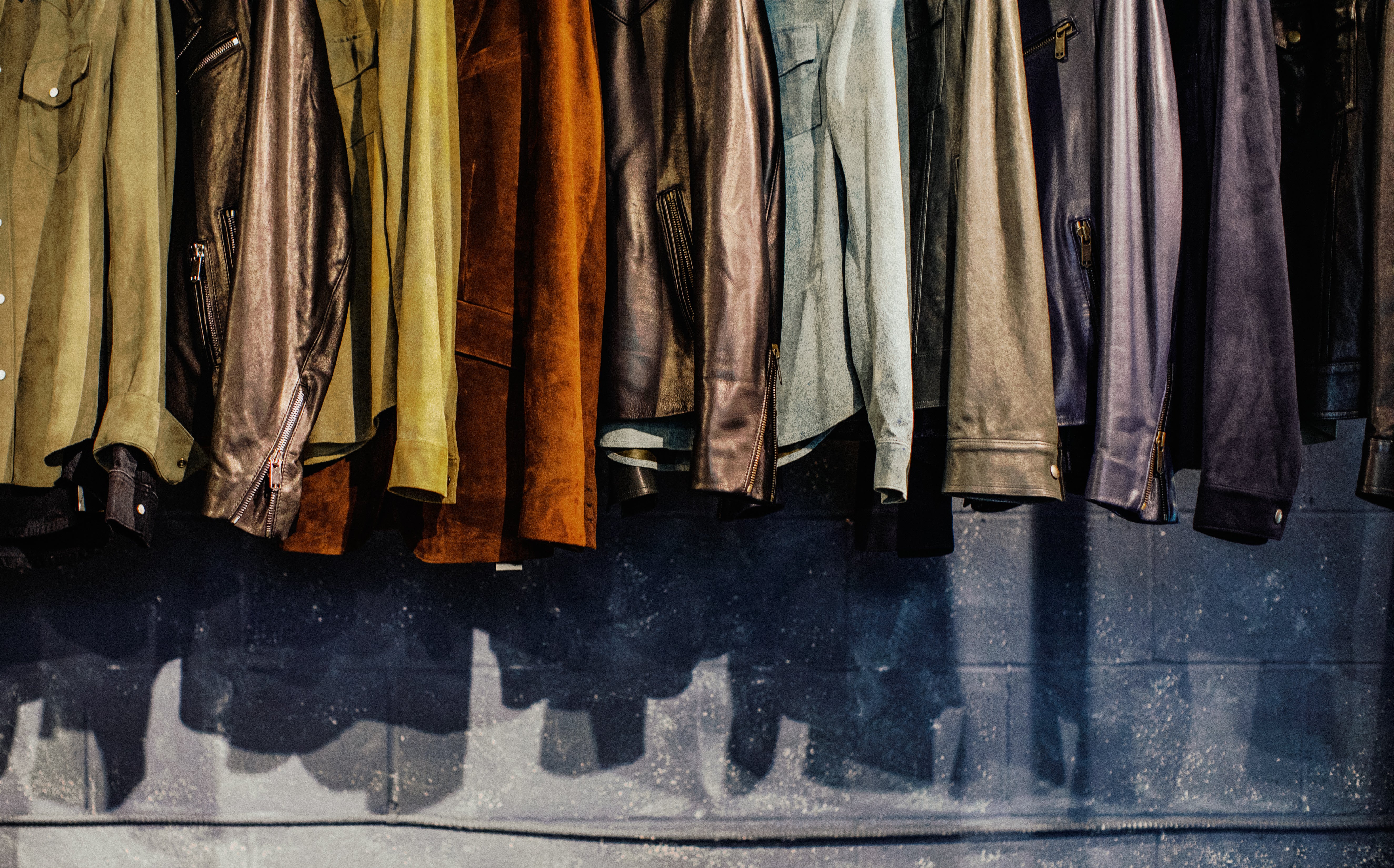 A row of leather and suede jackets and shirts hanging in various colors including brown, black and green, hanging against a dark blue wall with strong shadows.