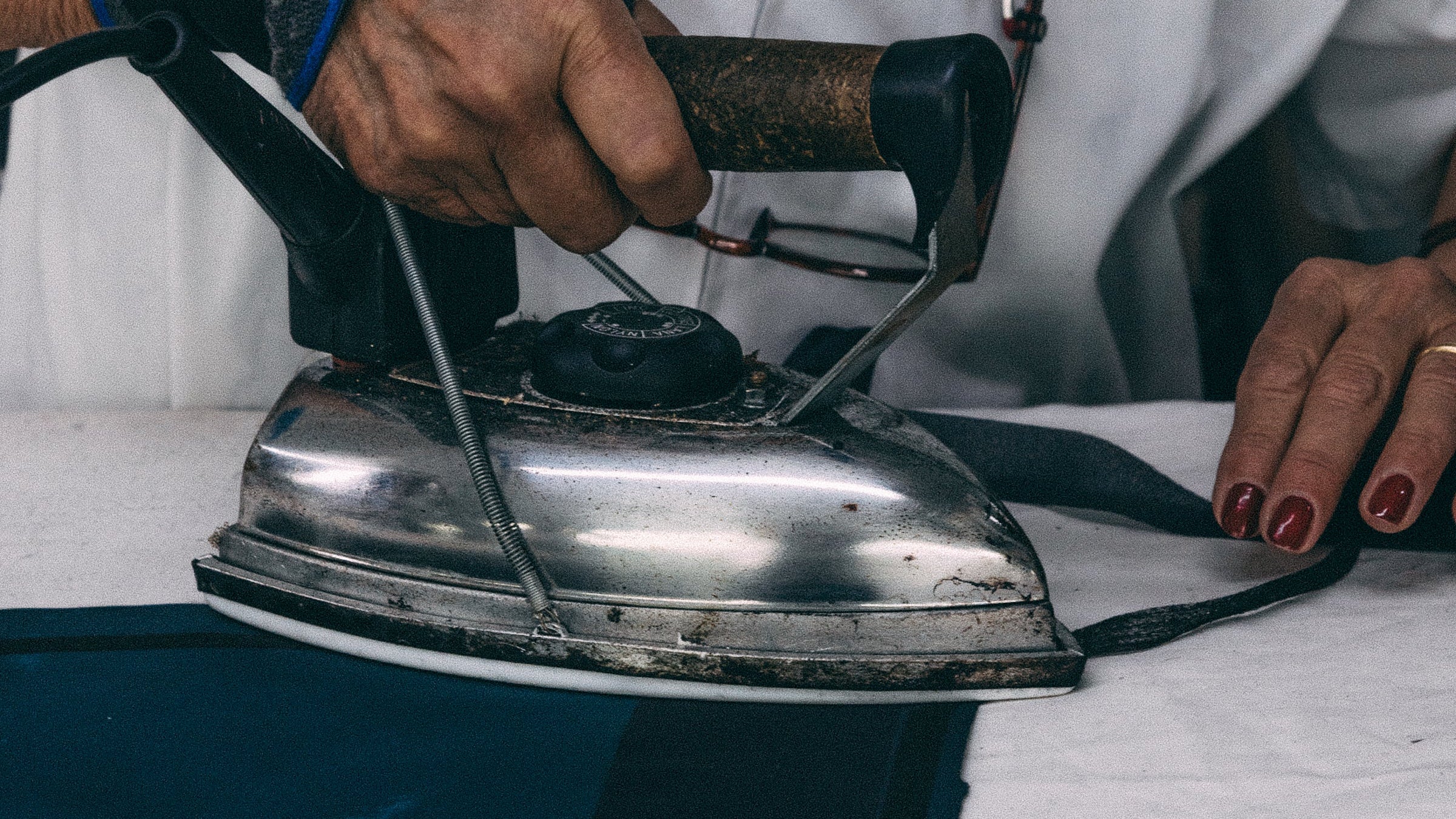 A close-up of a person in a white coat ironing a dark navy fabric with a vintage silver metal iron.